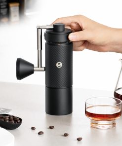 Hand holding the black Timemore S5 Pro grinder on a counter next to a glass of coffee