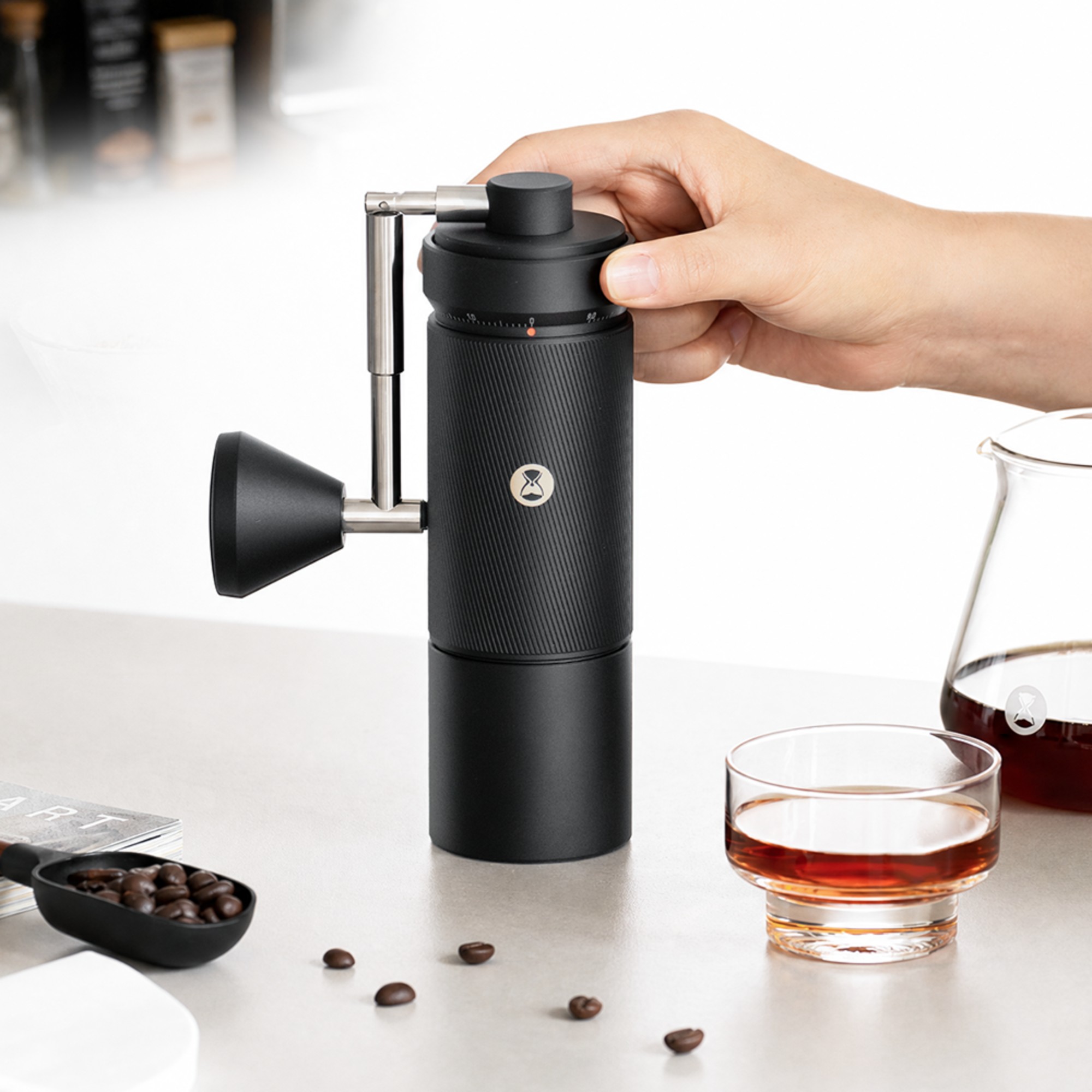 Hand holding the black Timemore S5 Pro grinder on a counter next to a glass of coffee